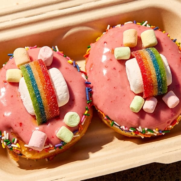 Two pink-frosted donuts with rainbow candy, marshmallows, and sprinkles in a takeout container.