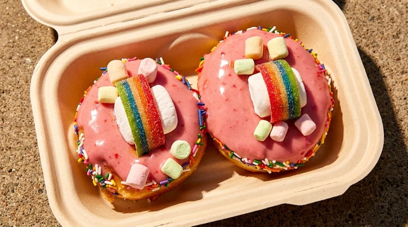 Two pink-frosted donuts with rainbow candy, marshmallows, and sprinkles in a takeout container.