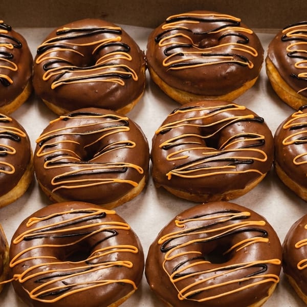 A dozen chocolate-frosted donuts with orange and black drizzle in a cardboard box.