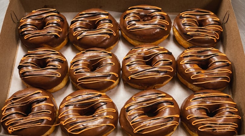 A dozen chocolate-frosted donuts with orange and black drizzle in a cardboard box.